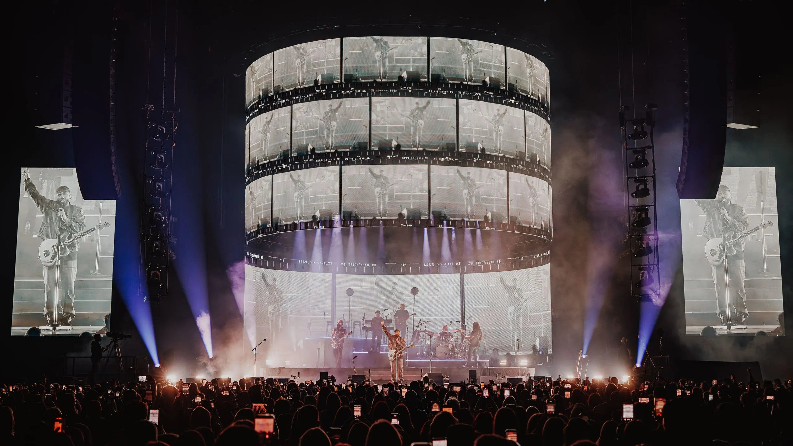 Cylindrical LED screen in three tiers of film-strip frames above the band, with portrait side screens and blue beams cutting through haze