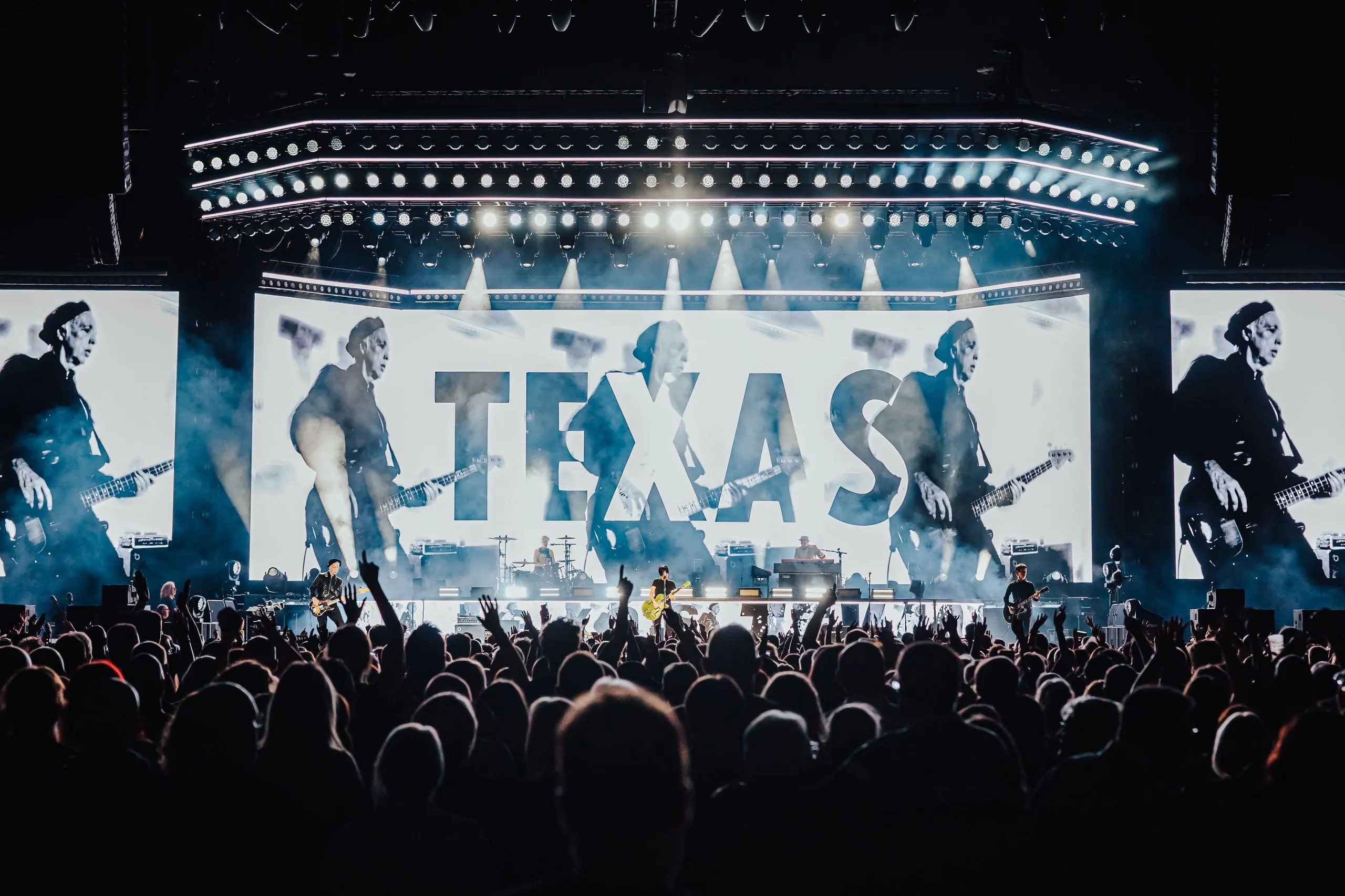 Band logo over black-and-white footage of the bass player on the wide LED wall, beneath a curved lighting rig, with the crowd's hands raised