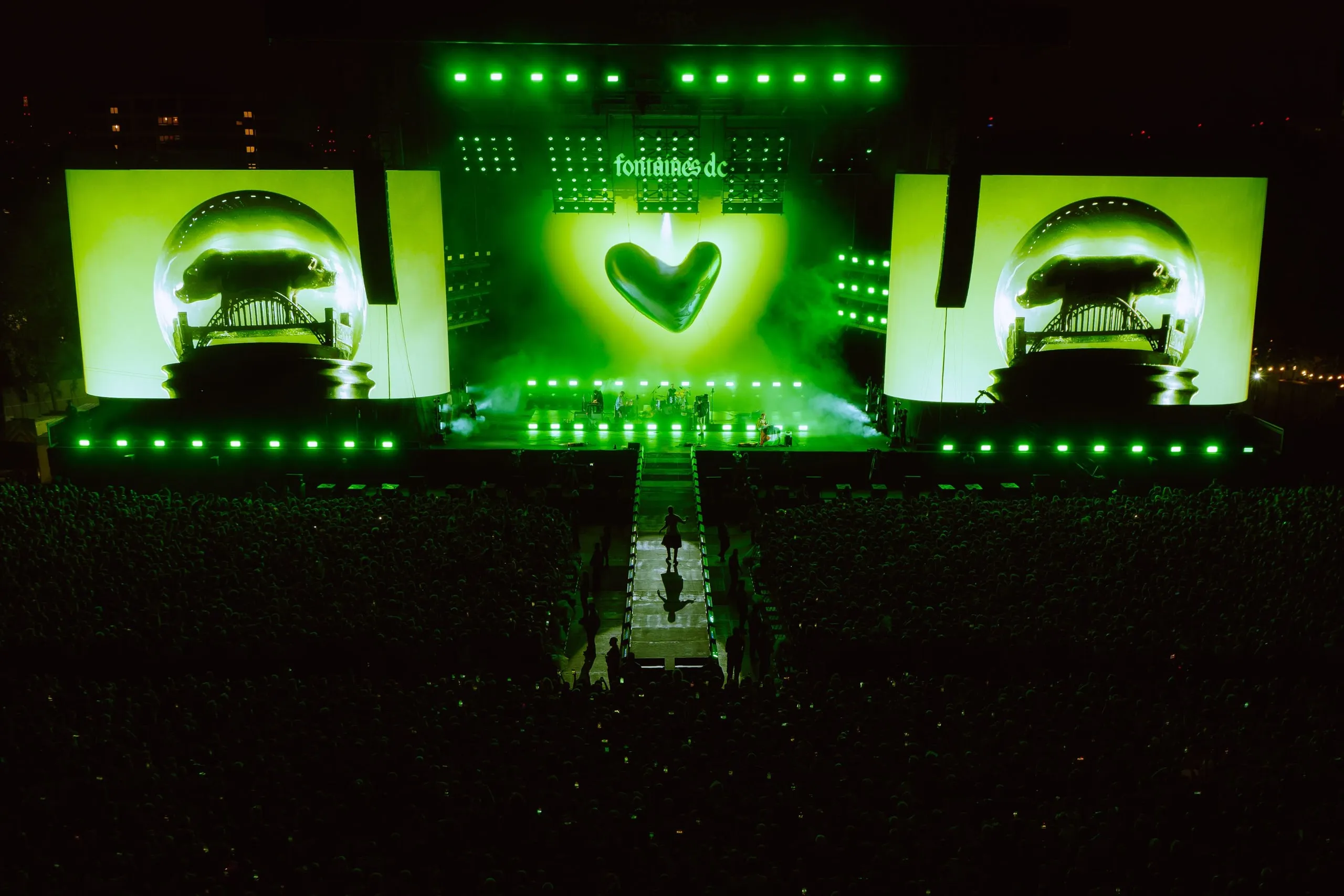 Green-lit stage at night with a heart on the centre screen and snow-globe graphics of a bridge on the side screens, a performer out on the catwalk