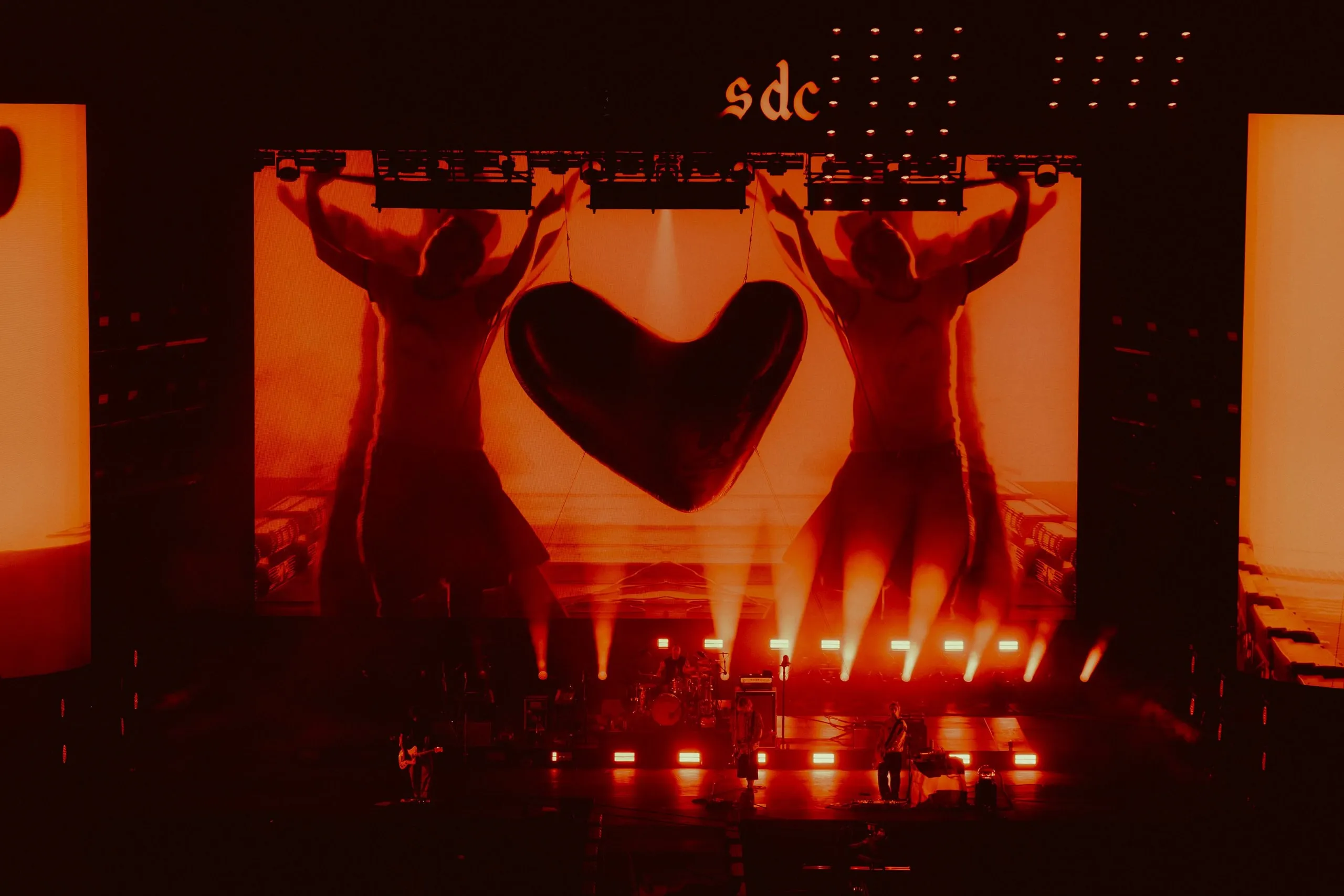 Hanging heart in front of an orange-red screen showing two mirrored figures, with the band in silhouette below