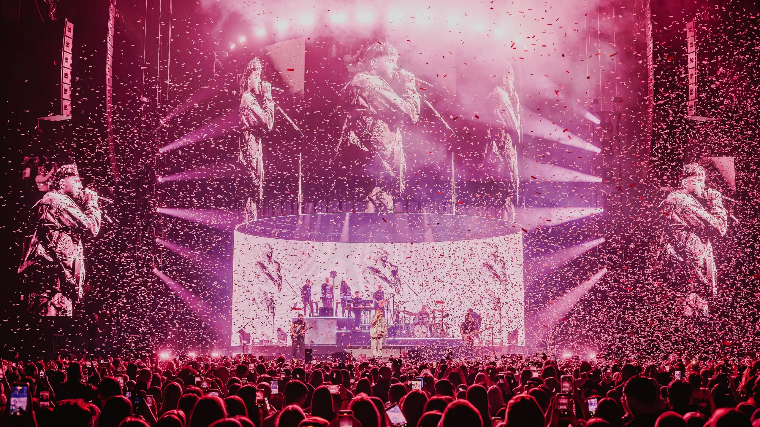 Arena filled with confetti under pink light, with performance footage across the screens and the band on stage below