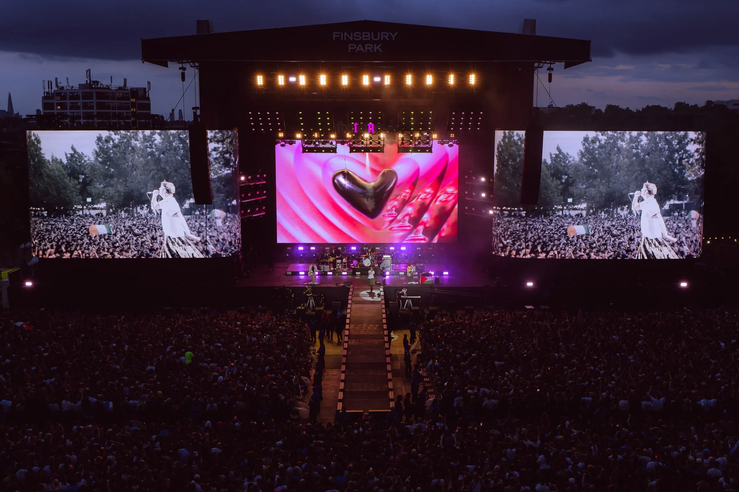 Finsbury Park at dusk, with a pink graphic and dark heart on the centre screen and the side screens showing the singer in front of the crowd