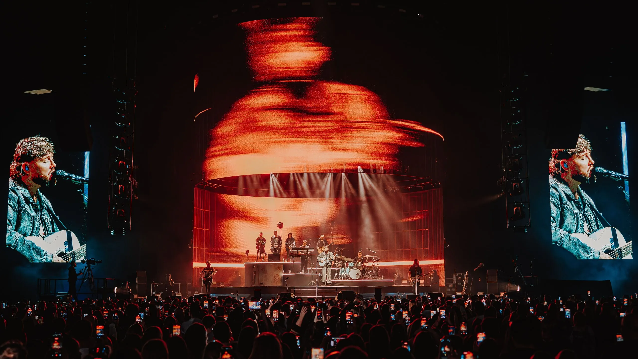 Blurred orange motion graphics on the cylinder screen while the side screens show close-ups of James Arthur singing with an acoustic guitar