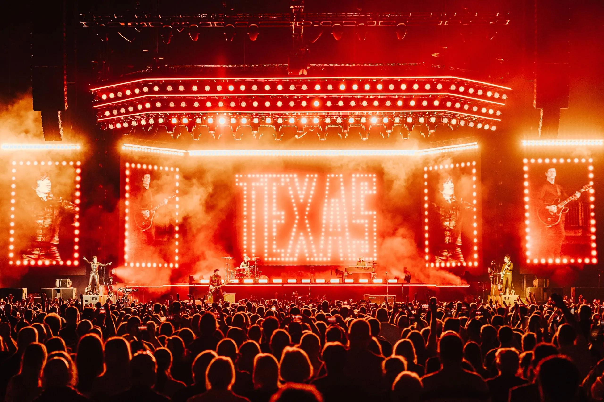 TEXAS spelled out in light bulbs on the centre screen, flanked by bulb-framed screens of the band, all in red light and haze