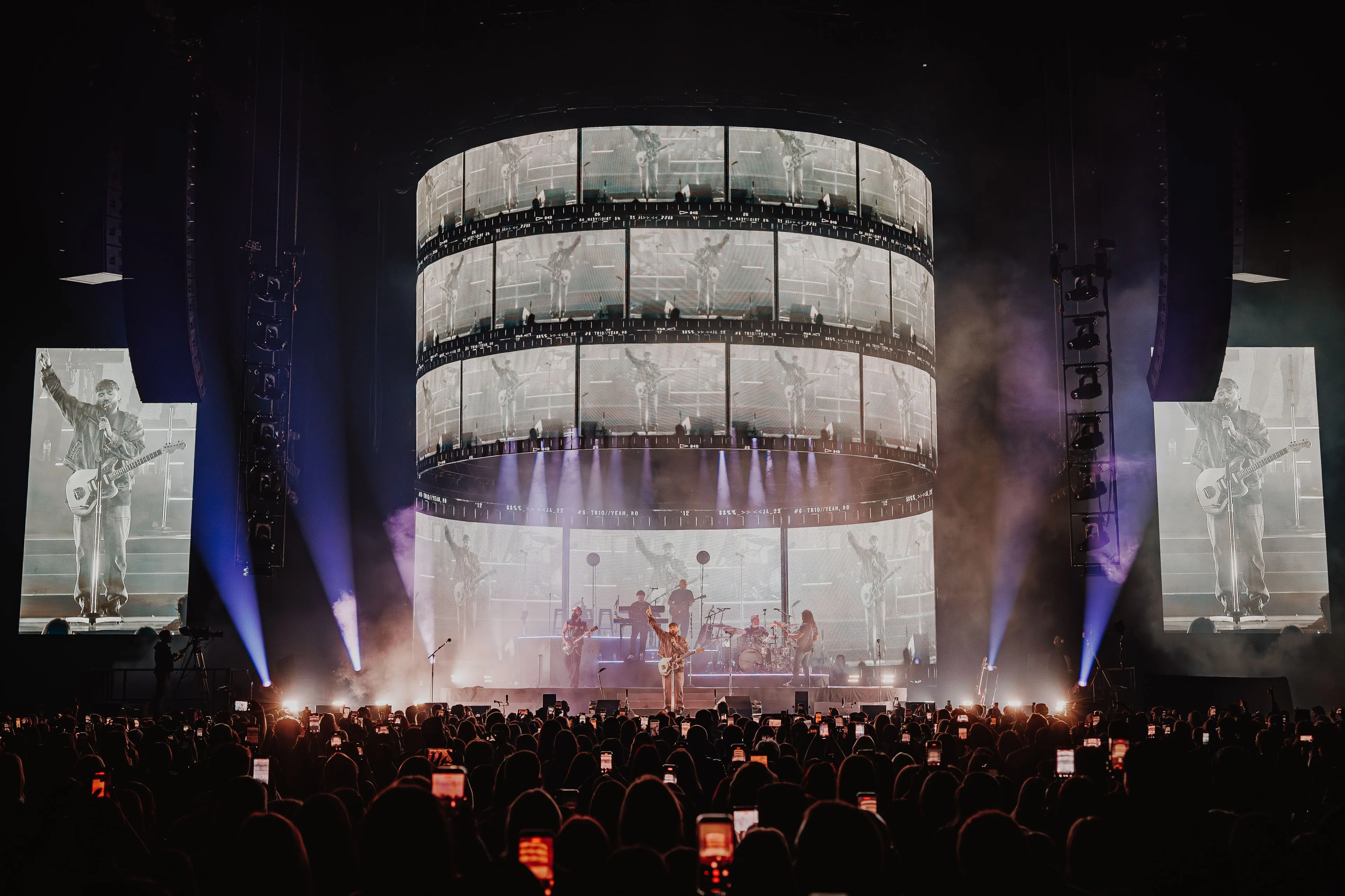 James Arthur performing beneath a tiered cylindrical LED screen of film-strip frames, with portrait side screens and an arena crowd holding up phones